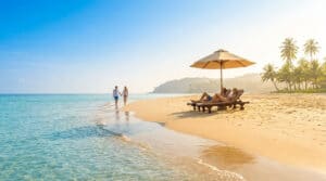 Un couple marche sur une plage paradisiaque alors qu'un autre se détend sous un parasol face à une mer turquoise.